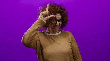 Elleriyle ezik bir jest yapan, gözlüklü ve kahverengi bir süveter giyen bir kadın, canlı mor bir arka plana karşı, duygularını ifade eden orta yaşlı, Hispanik bir kadına yer veriyor..