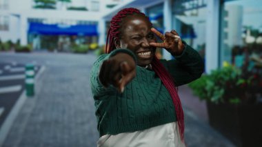 Şehir sokaklarında yeşil süveter ve pembe örgüler takmış, açık hava ortamında neşe ve çeşitliliği ifade eden barış işaretleriyle gülümseyen bir kadın..