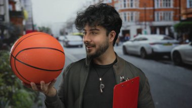 Sokakta basketbol ve pano tutan sakallı genç adam, sıradan bir hayat tarzı sergileyen kameraya hevesle gülümsüyor.