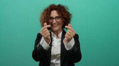 Yeşil arka planda kıvırcık saçlı, gözlüklü ve takım elbiseli bir kadın, kalp hareketi yaparken sevinç ifade ediyor..