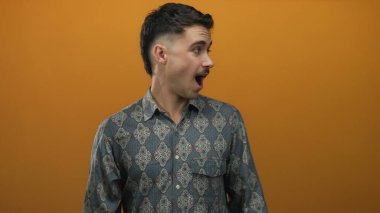 Şablonlu tişört giyen genç İspanyol adam canlı turuncu bir arka plana şaşırmış görünüyor, izole bir ortamda merak ve duygu yayıyor..