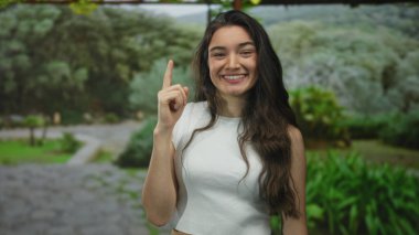 Açık havada güneşli yeşil bir parkta gökyüzüne parlak bir şekilde gülümserken işaret eden kadın iyimserlik.