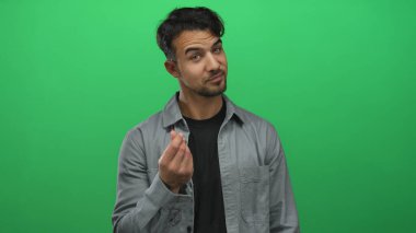 Genç İspanyol asıllı bir adam, yeşil bir arka plana parmaklarıyla para hareketi yapıyor. Günlük kıyafetleriyle neşeli ve kendinden emin bir tavır sergiliyor..