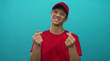 Kırmızı şapkalı ve gözlüklü genç İspanyol bir adam stüdyoda parmaklarıyla kalp hareketi yapar..
