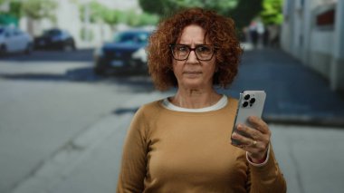 Şehir caddesinde duran bir kadın, elinde odaklanmış bir ifadeyle akıllı bir telefon, güneşli bir günde park etmiş arabalar ve yeşilliklerle çevrili..