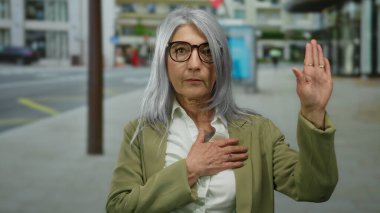 Gri saçlı yaşlı bir kadın şehir caddesinde, açık havada, gözlük takıyor, spor ceket giyiyor, şehir ortamında özgüven ve bağlılığı somutlaştırıyor..