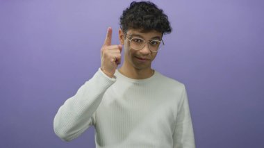 Gözlüklü ve beyaz kazaklı genç İspanyol adam mor stüdyoda kamerayı işaret ediyor..