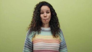 Kıvırcık saçlı İspanyol bir kadın, yumuşak sarı bir arka plana karşı neşeli bir balık yüzü takınıyor. Renkli bir kazak giyiyor..