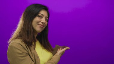 Sarı bluzlu İspanyol kadın, mor bir duvara karşı stüdyoda el ele sunarken gülümsüyor..