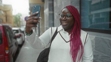 Pembe örgülü bir kadın arka planda arabaları olan bir caddede telefon görüşmesi yapıyor, akıllı telefon kullanıyor ve gülümsüyor..