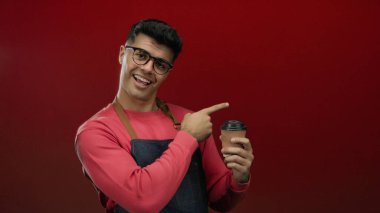 Gözlüklü ve önlüklü genç adam kırmızı arka planda kahve tutuyor, coşku ve güven ifadesiyle işaret ediyor..