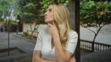 Güneşli bir parkta açık havada, yemyeşil giyinmiş, beyaz giyinmiş, düşünceli bir ifade takınan, sarı saçlar takınan, dingin bir ortam oluşturan bir kadın..