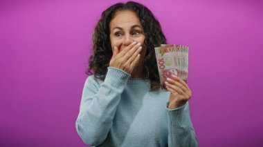 İzole edilmiş mor bir arka planda elinde yeni Zelanda doları tutan orta yaşlı bir kadın elinde para birimiyle şaşkınlık ve heyecan ifade ediyor..