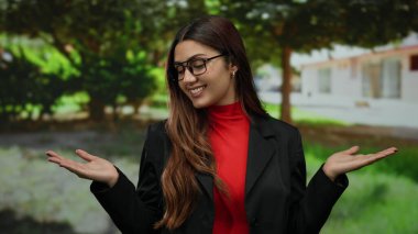 Parkta elleri açık, kırmızı ve siyah bir şekilde gülümseyen bir kadın, genç bir ifade, İspanyol, yeşil gözlüklü, etrafı yeşilliklerle çevrili..