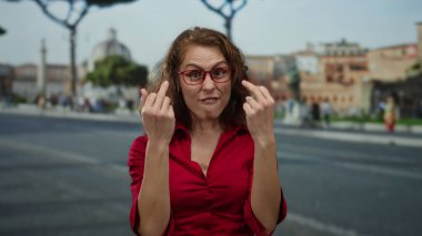 Kırmızı tişörtlü bir kadın, şehir sokaklarında tarihi binalarla anlamlı jestler yapıyor, açık havada duygusal bir tepki gösteriyor.