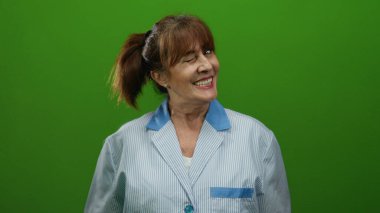 Kıdemli İspanyol kadın temizlik üniforması içinde canlı yeşil arka plana gülümsüyor, sıcaklığını ve dostluğunu profesyonel bir ortamda iletiyor..