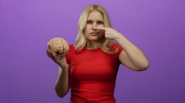 Kırmızı gömlekli kadın bir beyin modeli tutuyor ve mor bir arka plan karşısında dikilirken burnuna dokunuyor, odak ve konsantrasyon temalarını vurguluyor..