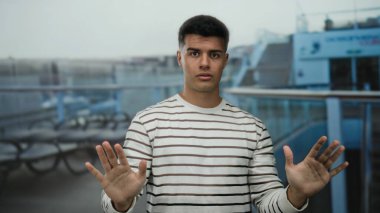 Bir yolcu gemisinin güvertesinde, elleri havada, çizgili gömlek giymiş, arka planda bulanık oturma ve gemi korkulukları olan bir adam..