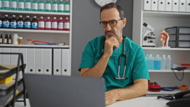 Klinikteki olgun İspanyol adam steteskoplu önlük giyiyor. Etrafı tıbbi malzemelerle çevrili. Profesyonel ortamdaki düşünceli konsantrasyonu yansıtıyor.