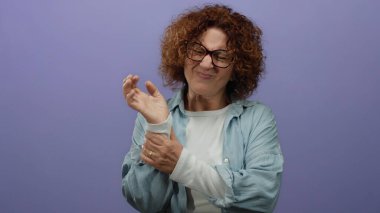 Mor duvarın önünde bileği ağrıyan bir kadın gözlük takıyor ve mavi bir gömlek giyiyor. Yüzünde endişeli bir ifade var. İçeriden izole edilmiş..