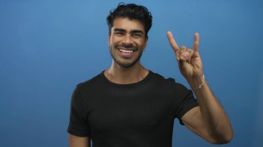 Genç İspanyol adam elinde kaya boynuzu hareketi yapıyor ve pürüzsüz mavi bir stüdyo duvarına geniş bir gülümseme; eğlence.