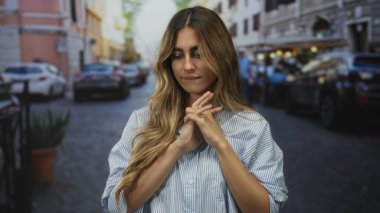 Sarışın kadın hareketli kaldırım taşı caddesinde dudaklara yakın ellerini çırpıyor; gerginlik beklentisi endişe yansıması.