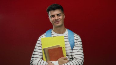 Sırt çantası ve not defterleriyle gülümseyen, parlak kırmızı arka planda, çizgili gömlek giyen, gündelik ve akademik bir stil sergileyen İspanyol genç adam..