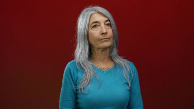 Gri saçlı yaşlı bir kadın canlı kırmızı bir arka plana karşı güvenle duruyor, bilgeliğini ve zarafetini basit turkuaz kıyafetleriyle sergiliyor, izole ve odaklanmış..