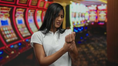 Meşgul bir kumarhanede bilek ağrısı çeken, arka planda renkli kumar makineleri olan bir kadın. Rahatsızlık ve ev ortamlarına odaklanıyor..