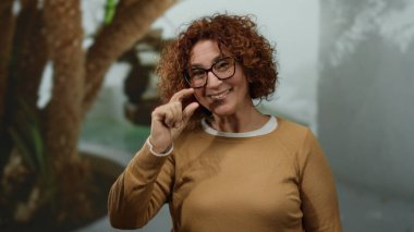 Bir kadın şehrin açık hava ortamında gülümsüyor ve el kol hareketi yapıyor kıvırcık saçlarını ve gözlüklerini parlaklaştırıyor, anlamlı ve neşeli bir anı yakalıyor..