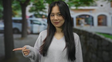 Uzun saçlı, gülümseyen ve ağaçlarla çevrili bir caddeyi işaret eden, arka planı bulanık, açık hava manzarasını çağrıştıran genç bir kadın..
