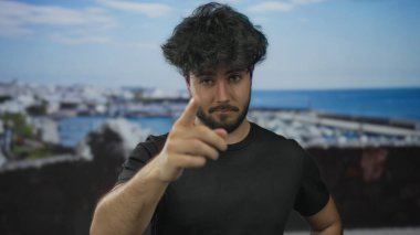 Sakallı genç adam limana yakın bir yerde duruyor. Arkaplanı açık gökyüzünün altında, gündüzleri siyah tişört giyerken.