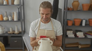 Uzun saçlı, önlüklü sarışın bir adam stüdyoya bakarken kırmızı akıllı telefon tutuyor..