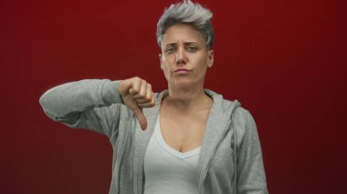 Kırmızı stüdyoda gri kapüşonlu kısa gri saçlı bir kadın baş parmak hareketi gösteriyor; reddedilme hüsranı.