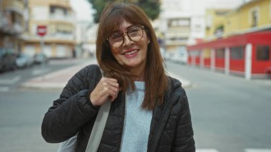 Şehrin sokak ortamında güvenle gülümseyen İspanyol asıllı son sınıf öğrencisi kadın, şehir arka planında, açık havada yakalanmış, açık renk süveter, gözlük ve gündelik ceket, somut modern yaşam tarzı..