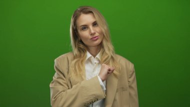 Yeşil ekranın önünde bej renkli ceketli bir kadın. Güvenle poz veriyor. Yumruğunu kaldırmış, güç ve kararlılığını gösteriyor..