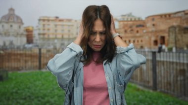 Latin kadın, Roma harabeleri yakınlarındaki gürültü nedeniyle kulaklarını kapatıyor. İfadesi, tarihi şehir zeminine karşı rahatsızlığını gösteriyor ve kentsel zorlukları vurguluyor..