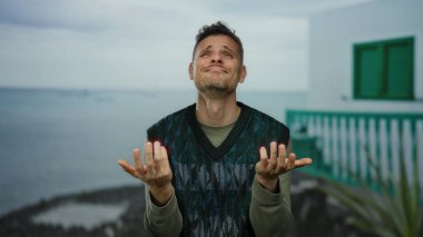 Okyanus kıyısında gezinen ve bulutlu gökyüzünün altında gelişigüzel inşa edilen duygusal ifadeli genç adam..