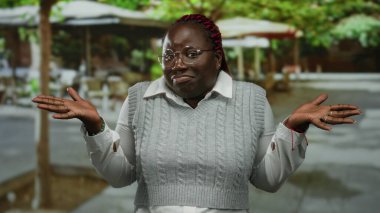 Örgülü saçlı ve gözlüklü bir kadın şehir kafesindeki terasta omuz silkiyor, beyaz gömleğin üzerine gri bir kazak giyiyor, sokak ortamında sıradan bir ifadeyi somutlaştırıyor..