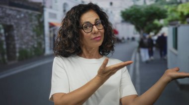 Orta yaşlı İspanyol kadın, bir şehir caddesinde ürün sunarken ellerini açık gösterdi; onay iyimserliği.