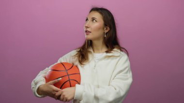 İzole edilmiş pembe arka plana karşı düşünceli bir ifadeyle basketbolu tutan kadın dinamik poz ve renkli kontrastla atletik yaşam tarzını yakalıyor..
