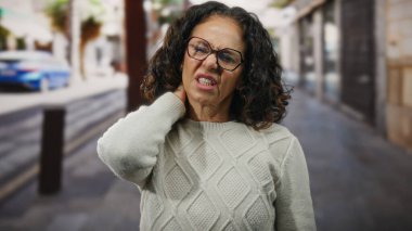 Kazak giyen kıvırcık saçlı bir kadın şehir sokaklarında boynu ağrıyor..