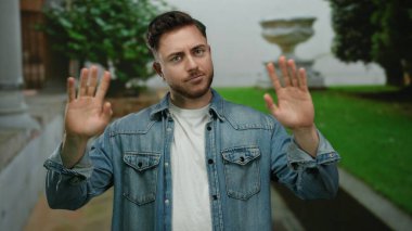 Sakallı genç adam şehir dışında bir sokak ortamında elini kaldırıyor, kot ceket giyiyor, düşünceli bir bakışla jest yapıyor, yemyeşil ve şehir mimarisi ile çevrili..