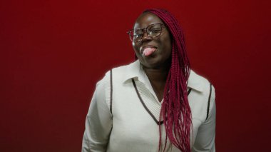 Gözlüklü ve uzun örgülü Afro-Amerikan bir kadın neşeli kırmızı bir arka plana karşı dilini şakayla uzatıyor, şık bir elbiseyle neşe ve eğlenceyi ifade ediyor..