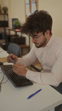 Ofiste akıllı telefon kullanan, klavye, gözlük, kağıt ve kalemle masa başında çalışan, kapalı alanda verimli bir sahne yaratan sakallı genç adam..