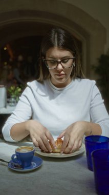 Güneşli bir Akdeniz restoranında hamur işinden hoşlanan, gözlüklü ve beyaz gömlekli, mavi bardaklarla çevrili ve masanın üzerinde küçük bir kahve olan bir kadın, sıcak bir ortam yaratıyor..