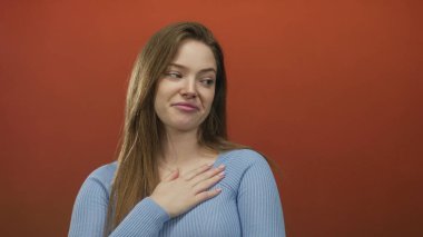 Stüdyoda mavi süveter giyen bir kadın kırmızı arka plana sakin bir tavırla bakıyor; belirsizlik..