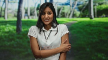 Beyaz gömlekli, yaka kartı takmış, arka planda yemyeşil bir çevre ve ağaçların arasında düşünceli bir şekilde duran genç bir kadın..