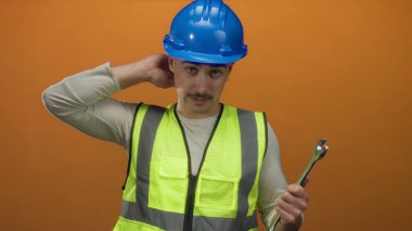 Yansıtıcı yelekli ve kasklı genç adam turuncu arka planda İngiliz anahtarı tutuyor, düşünceli görünüyor..