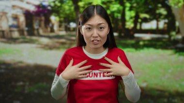 Güneşli bir günde, Çin 'de bir parkta gönüllü olarak çalışan bir kadın kırmızı bir gömlek giyiyor ve yeşil yemyeşil bir zemine karşı neşeyle gülümsüyor, neşe ve coşku ifade ediyor..